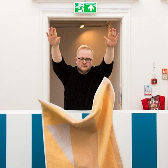 A person dressed in a black jumper throws a yellow piece of fabric over a blue and white wall. They are stood in front of a doorway.