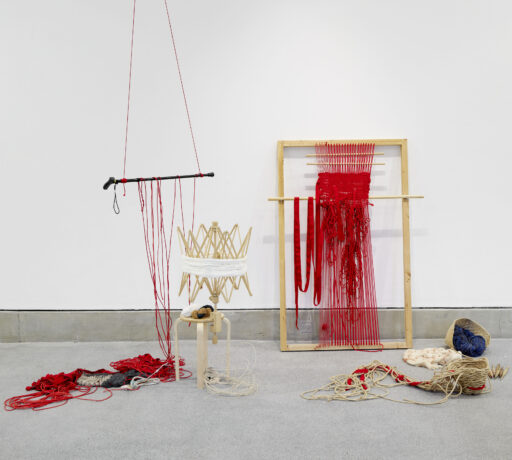Image shows Raisa Kabir's art instillation. A wooden structure with red cloth, hanging down. To the left is a walking stick suspended by red cloth and ropes run along the floor.